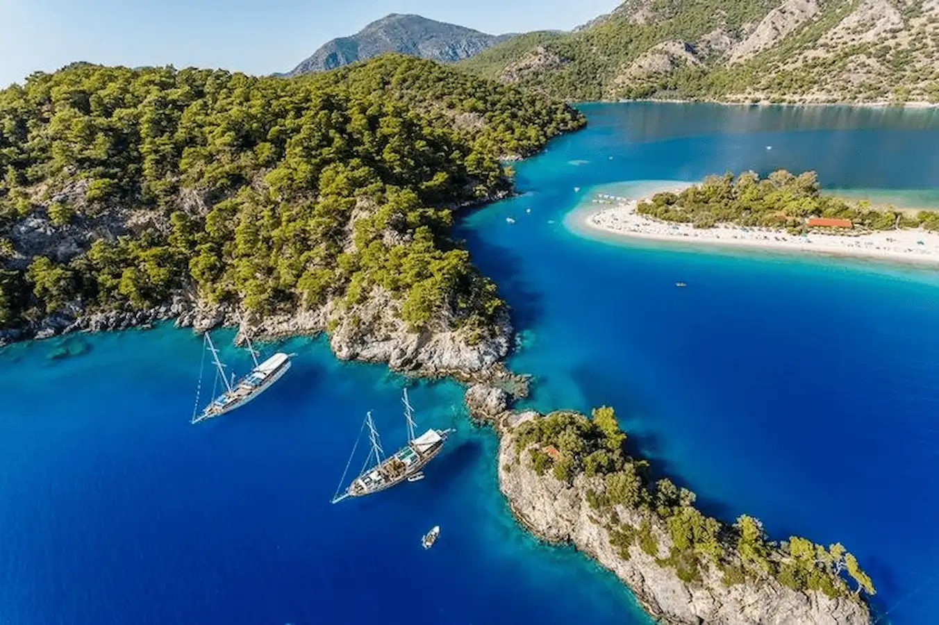 A sailing yacht and a classic wooden gulet drifting on turquoise waters along the rugged Turkish coastline.