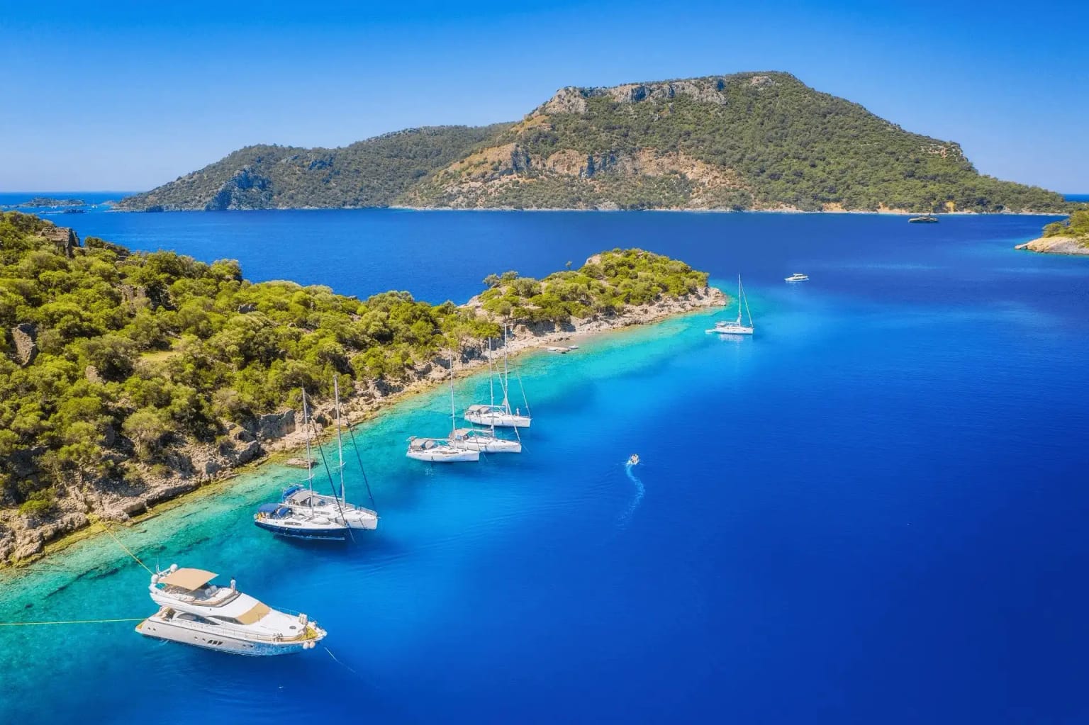 Eine Gulet, die in einer türkisfarbenen Bucht an der Ägäis- und Mittelmeerküste vor Anker liegt, jemand beim Paddeln, eine Segelyacht und ein Katamaran.